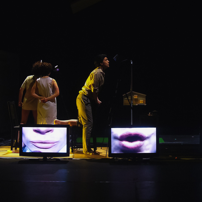 Three performers in 1950's costume stood at microphones behind a row of TV screens showing just their mouths (Virtuoso).