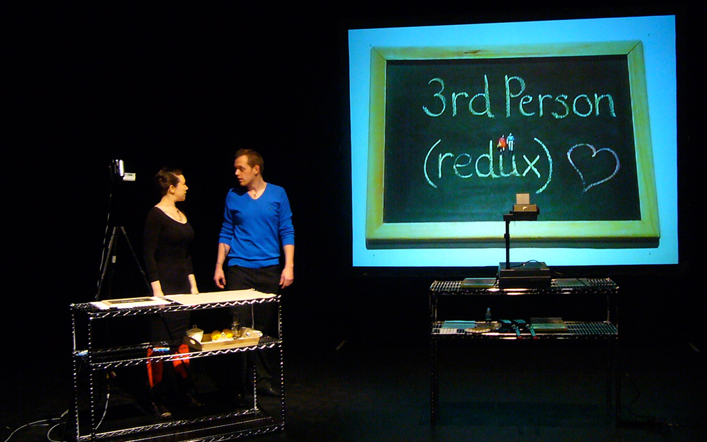 Gillian Lees and Andrew Westerside stood behind a counter, on stage. An image of a blackboard is projected on the wall which reads "3rd Person (redux) *heart*" (Third Person).