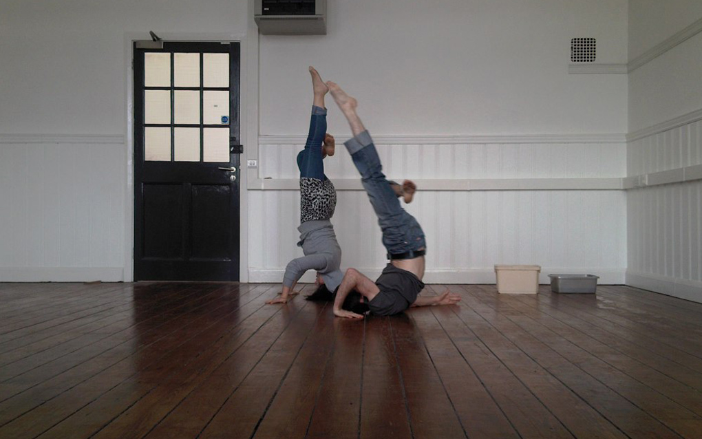 Two participants of a Proto-type Summer School, performing head and shoulder stands in an empty room with a wooden floor.