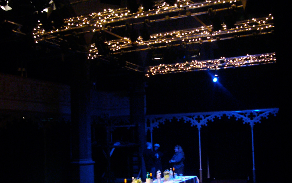 A long dining table, laid for dinner, on a stage, under fairy lighting.