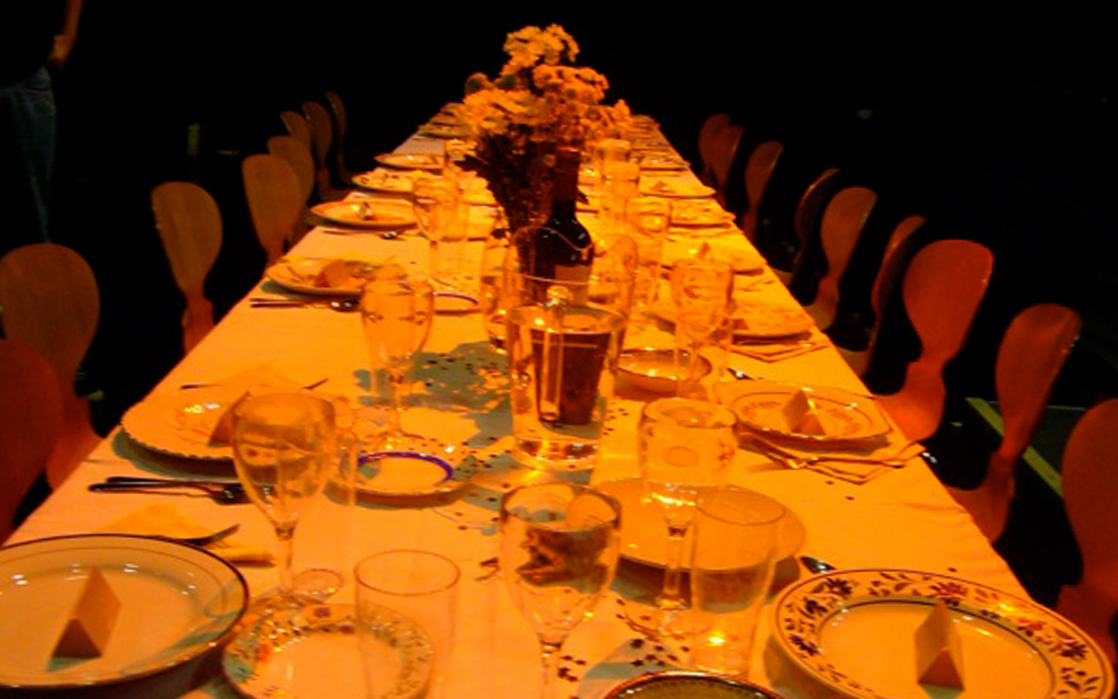A long dinner table, laid with mis-matched vintage crockery, on a stage in a theatre.