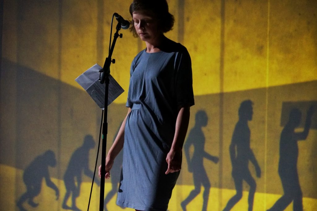Performer Leentje Van de Cruys wearing a grey dress, stood at a microphone (The Good, the God, and the Guillotine).