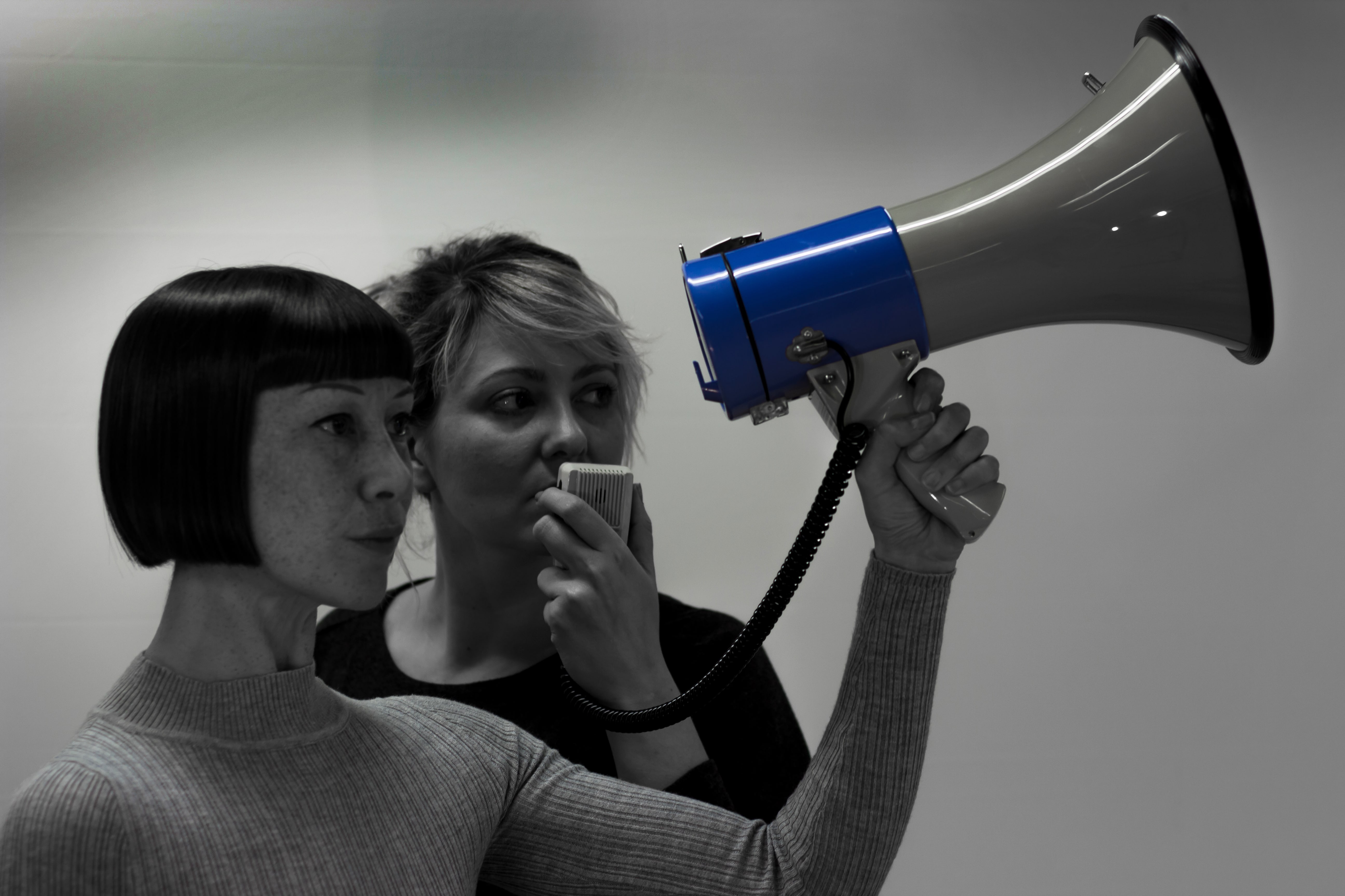 Main promo image for Proto-type Theater's 'The Audit (or Iceland, a modern myth)'. Two women stand side-by-side speaking into a blue loudhailer..