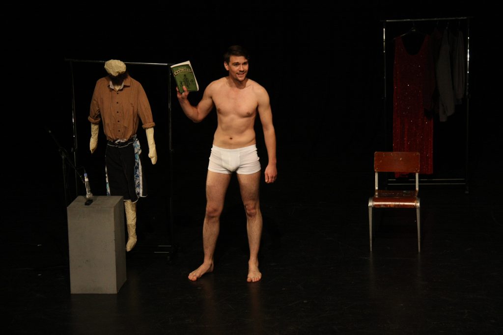 Jon from Sticky Tape Theatre, stood on stage, wearing pants, holding a book